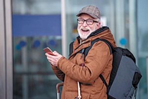 Laughing old gray-haired man with beard is standing outdoors with backpack and mobile phone. He is looking at camera with wide smile